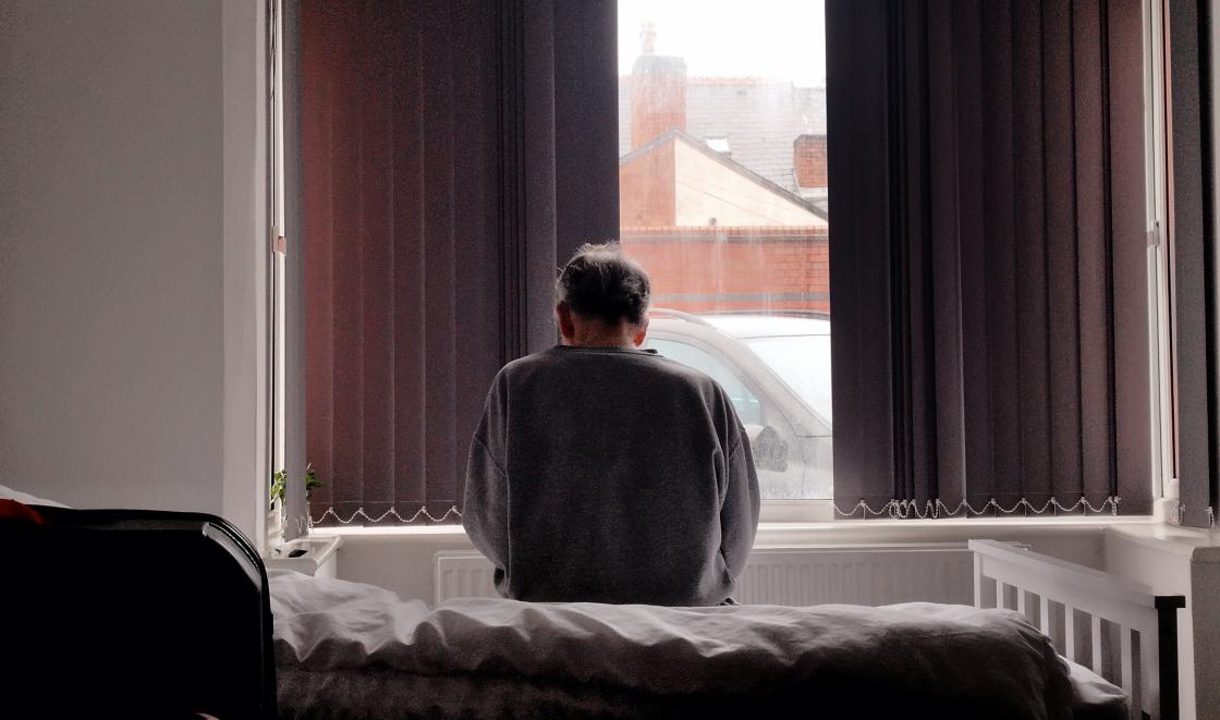 An older man sitting on a bed facing a window, with curtains drawn halfway across, suggestive of loneliness and isolation 